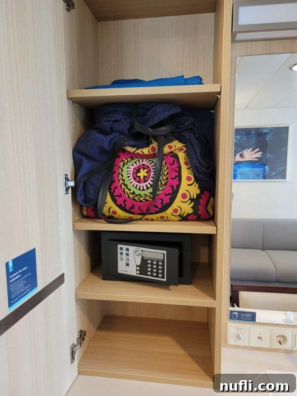 A compact cabinet with a small safe, a vibrant colorful bag, and a neatly folded sweatshirt, illustrating efficient storage solutions within a cruise ship stateroom.