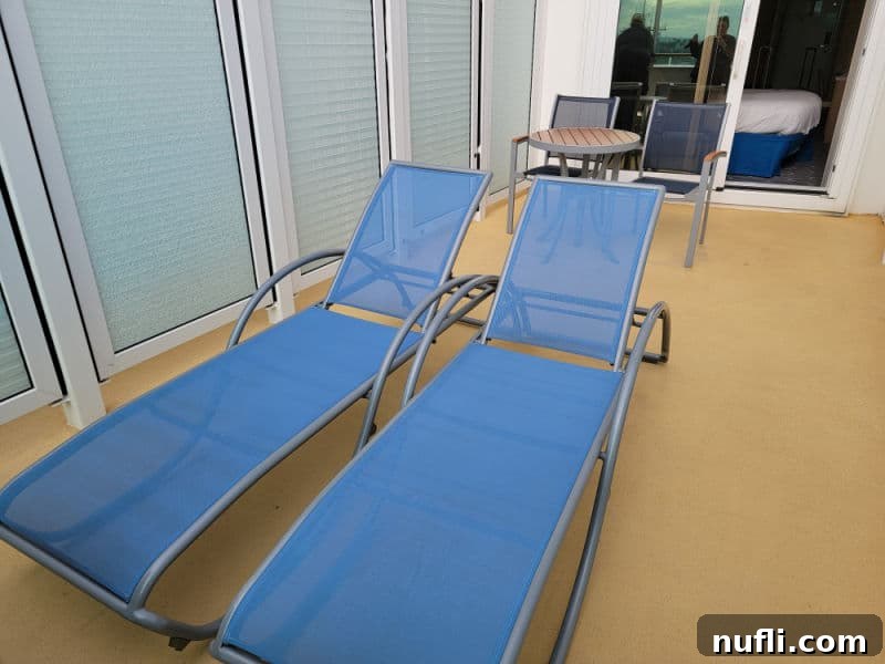 Two luxurious lounge chairs grace an extended cruise ship balcony, with a table and chair positioned just behind them. An open sliding glass door offers a peek into the stateroom's inviting interior.