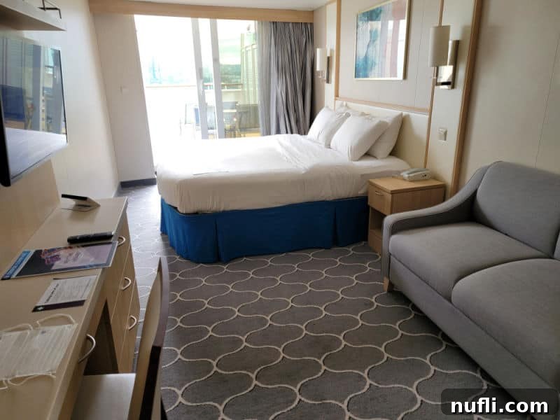 A inviting cruise ship stateroom, showcasing a comfortable grey couch, a plush bed, an open sliding glass door leading to a balcony, and a cabinet along one wall topped with a flat-screen TV.