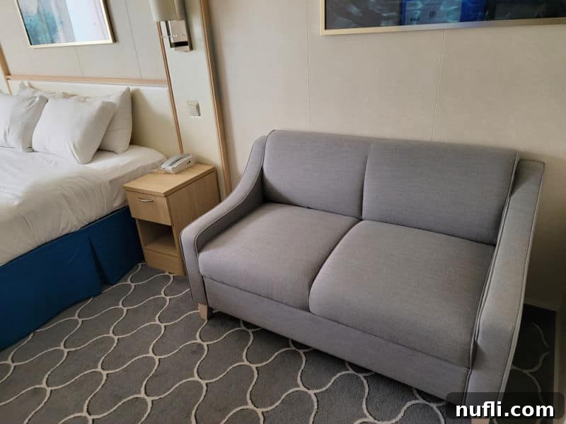A cruise ship stateroom featuring a small grey couch for two, positioned alongside a comfortable bed and a functional nightstand, creating a cozy and efficient living space.