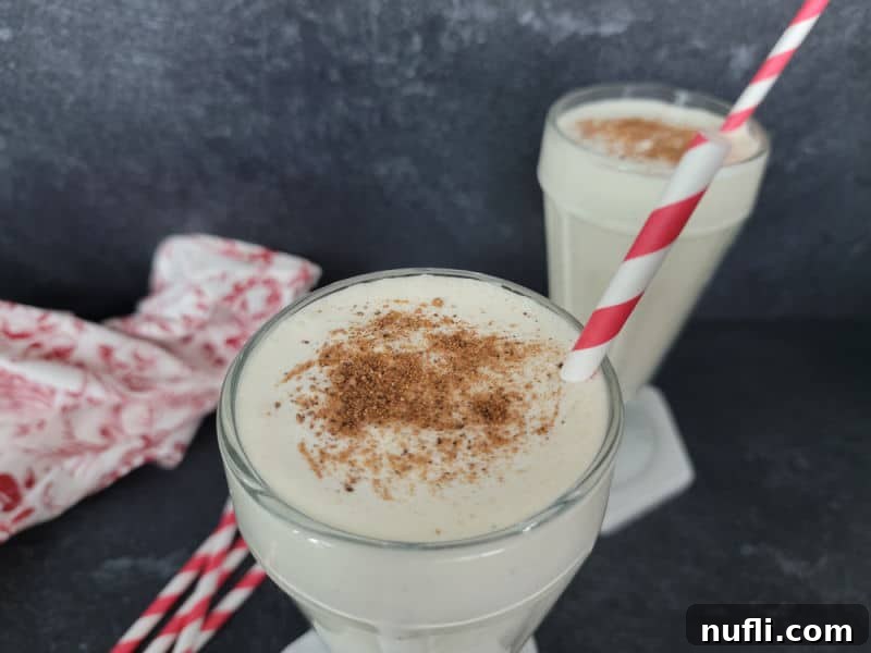A freshly blended eggnog shake in a milkshake glass, topped with a dusting of ground nutmeg and featuring a festive red striped straw, ready for sipping.
