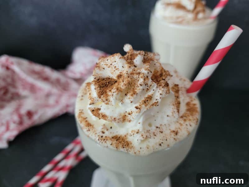 A beautifully presented Eggnog milkshake in a glass, generously garnished with whipped cream and freshly ground nutmeg, accompanied by a vibrant red striped straw.