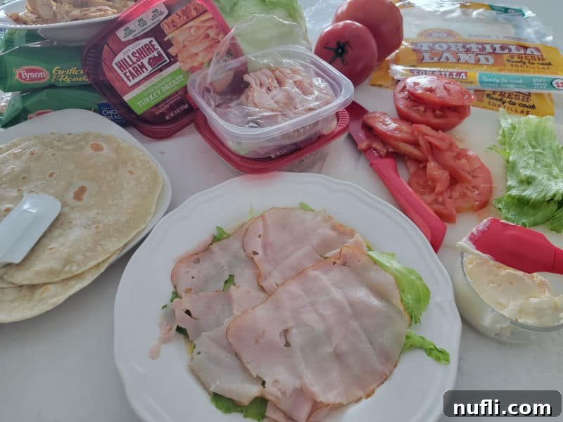 ham on lettuce and a tortilla next to a plate of tortillas, lettuce, tomatoes, mayo, and a knife