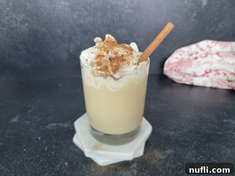 Decadent Eggnog Mudslide 8 A clear glass with an Eggnog Mudslide, elegantly topped with whipped cream, a dusting of ground cinnamon and nutmeg, and a decorative cinnamon stick for garnish.