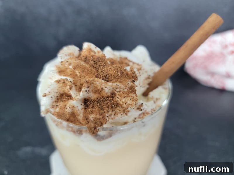 Decadent Eggnog Mudslide 9 Close-up of an Eggnog Mudslide cocktail topped with whipped cream, generously sprinkled with ground nutmeg, and garnished with a cinnamon stick in a clear glass.