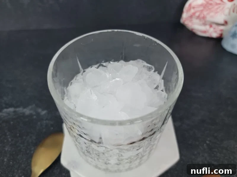 A clear crystal glass filled with ice cubes, positioned beside a gleaming gold spoon, ready for cocktail preparation.