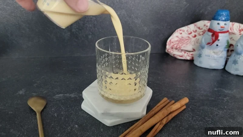 Rich, creamy eggnog being poured into a chilled glass, with a few cinnamon sticks and a spoon nearby, setting the stage for a festive drink.