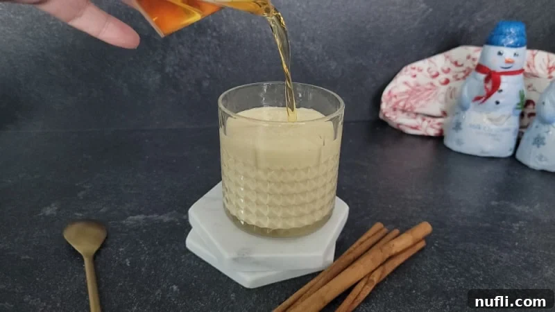 The fiery amber stream of Fireball Cinnamon Whiskey being added to the creamy eggnog in a glass, with cinnamon sticks and a spoon waiting beside it.
