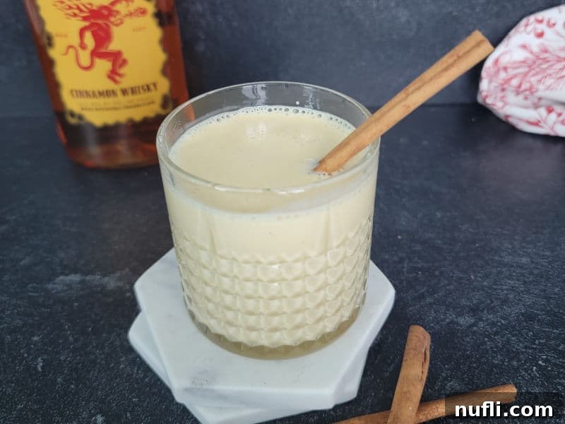 A festive glass of Fireball Eggnog, garnished with a single cinnamon stick, with the Fireball bottle positioned behind it on a rustic surface.