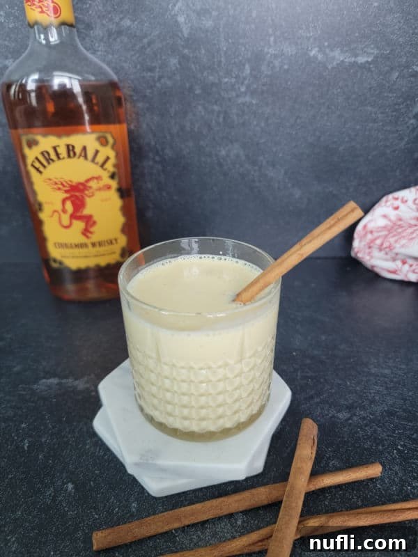 A festive glass of Fireball Eggnog on a white coaster, adorned with cinnamon sticks, positioned in front of a bottle of Fireball Cinnamon Whisky, creating a cozy holiday vibe.