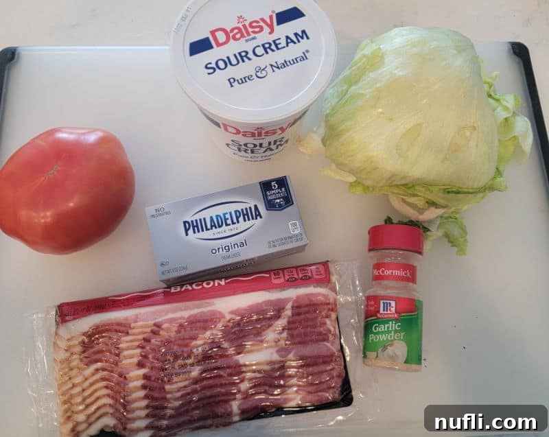 Ingredients for BLT Dip: sour cream, a head of fresh lettuce, ripe tomato, cream cheese, a package of bacon, and garlic powder, arranged on a rustic cutting board.