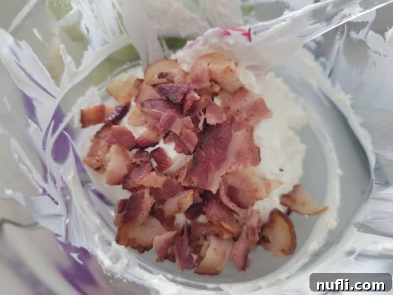 Chopped crispy bacon pieces gently folded into the smooth cream cheese mixture in a glass bowl, with a spatula demonstrating the mixing process.
