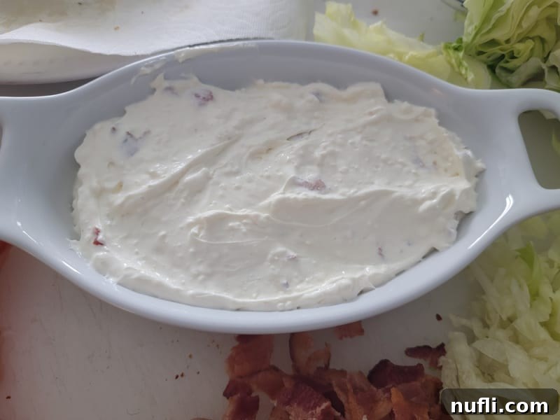 The creamy dip mixture in a white serving bowl, with separate piles of cooked bacon and shredded lettuce next to it, awaiting final assembly.