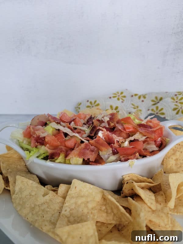 A white serving bowl filled with a beautifully garnished BLT Dip, topped with crispy bacon, shredded lettuce, and diced tomatoes. Tortilla chips and a decorative cloth napkin are arranged nearby.