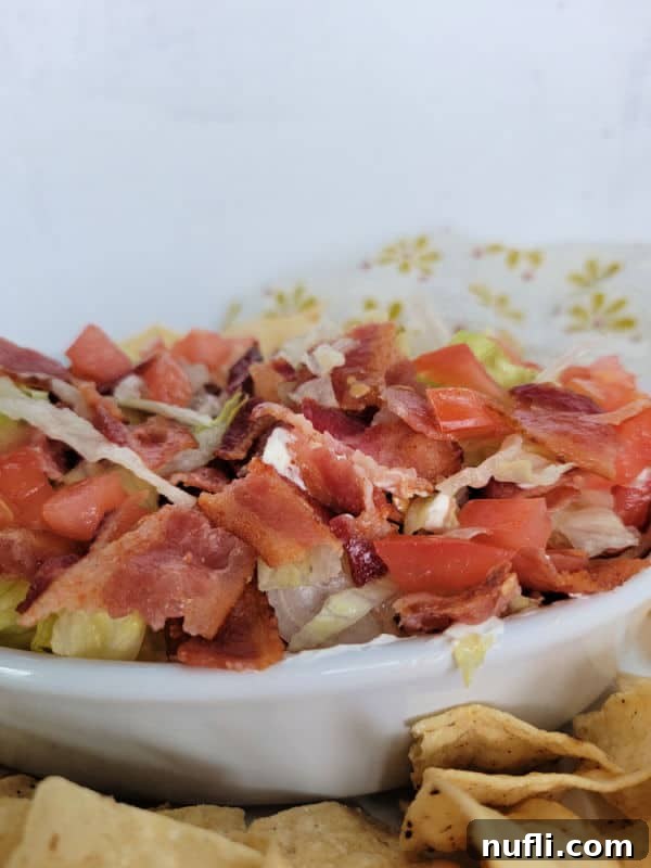 A close-up of BLT Dip in a white bowl, showing the texture of the creamy base and the fresh toppings: crispy bacon, vibrant diced tomatoes, and green shredded lettuce, with tortilla chips nearby.