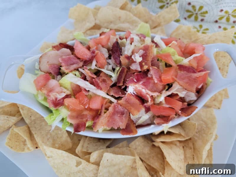 A top-down view of BLT Dip in a white bowl, generously topped with diced bacon, tomatoes, and shredded lettuce, accompanied by a scattering of sturdy tortilla chips.
