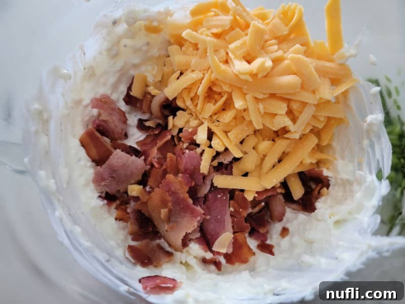 Chopped bacon and shredded cheddar cheese sprinkled over the creamy sour cream and ranch mixture in a glass bowl, ready to be folded in.