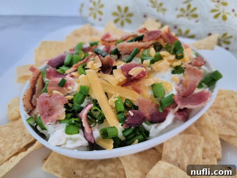 A large white bowl brimming with Bacon Cheddar Ranch Dip, garnished with extra bacon and green onions, surrounded by a generous spread of crispy tortilla chips.