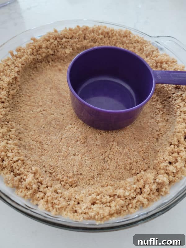Graham Cracker Crust in a glass pie pan with a purple measuring cup pressing graham cracker crumb mixture
