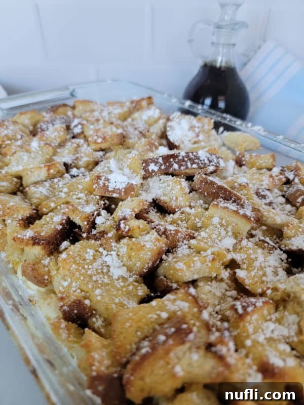 A beautiful shot of the overnight French Toast breakfast casserole in a baking dish, perfectly golden, with a jar of rich maple syrup positioned invitingly beside it.