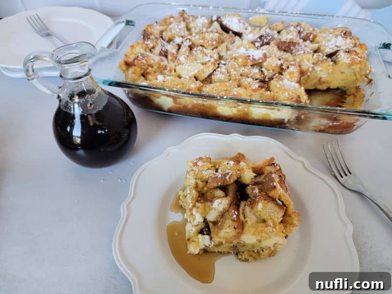 A freshly baked Overnight French Toast casserole in a glass baking dish, with a single, perfectly cut piece placed on a white plate with a fork, and a bottle of maple syrup nearby.
