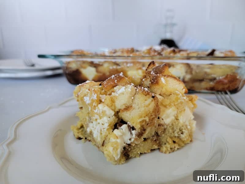 A delicious piece of Overnight French Toast breakfast casserole on a pristine white plate with a fork, perfectly positioned in front of a full baking dish of the casserole.