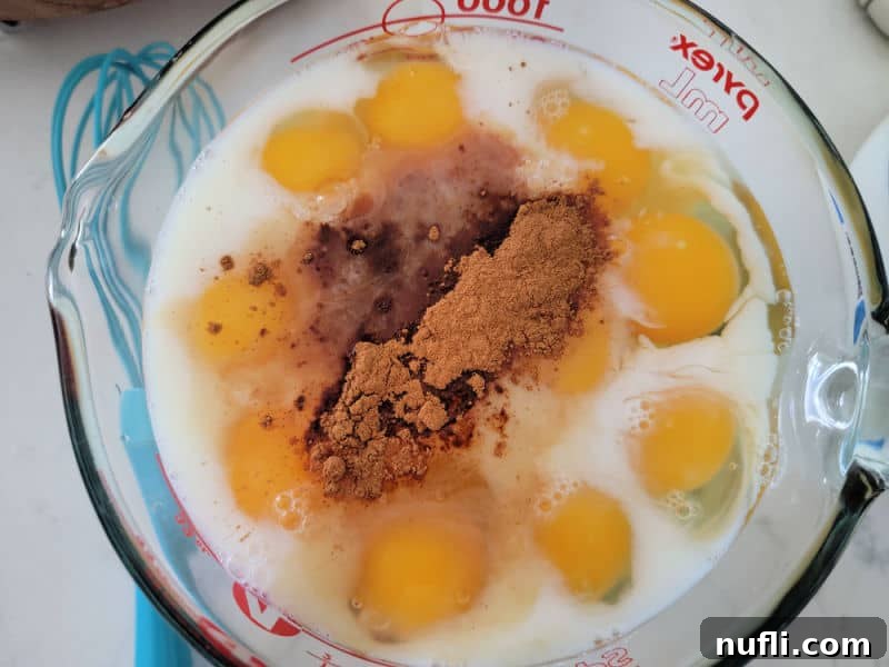 Close-up of a large Pyrex measuring bowl with a golden egg, milk, and ground cinnamon mixture, next to a blue whisk, for the French Toast Overnight Casserole custard.