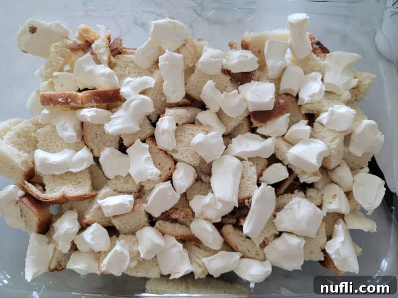 Small cubes of cream cheese distributed over the first layer of bread in a casserole dish, preparing for the French Toast overnight breakfast casserole.
