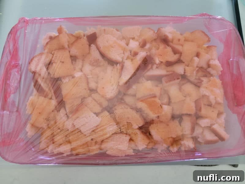 A glass casserole dish filled with the prepared French Toast overnight breakfast casserole, tightly covered with vibrant red plastic wrap, ready for refrigeration.