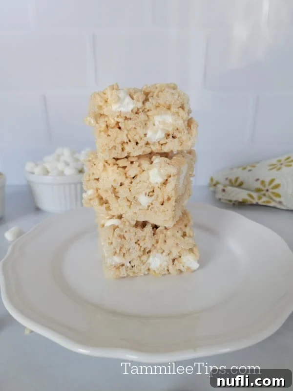 Stack of 3 Rice Krispie Treats on a white plate with mini marshmallows in a bowl behind 