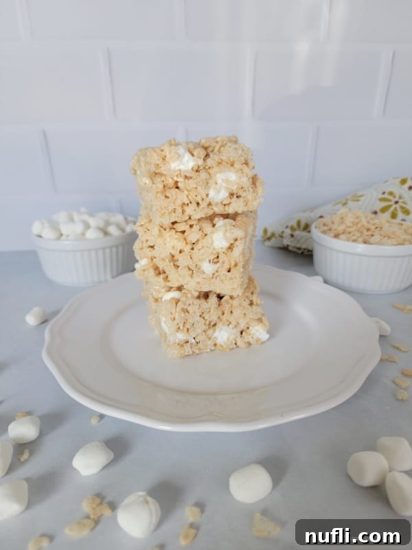 Stack of three Rice Krispie Treats on a white plate with mini marshmallows and rice cereal surrounding the plate