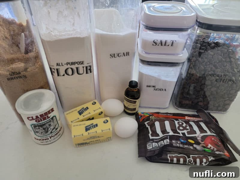 Baking ingredients including brown sugar, flour, granulated sugar, salt, baking powder, butter, eggs, chocolate chips, and an M&M package on a white counter.