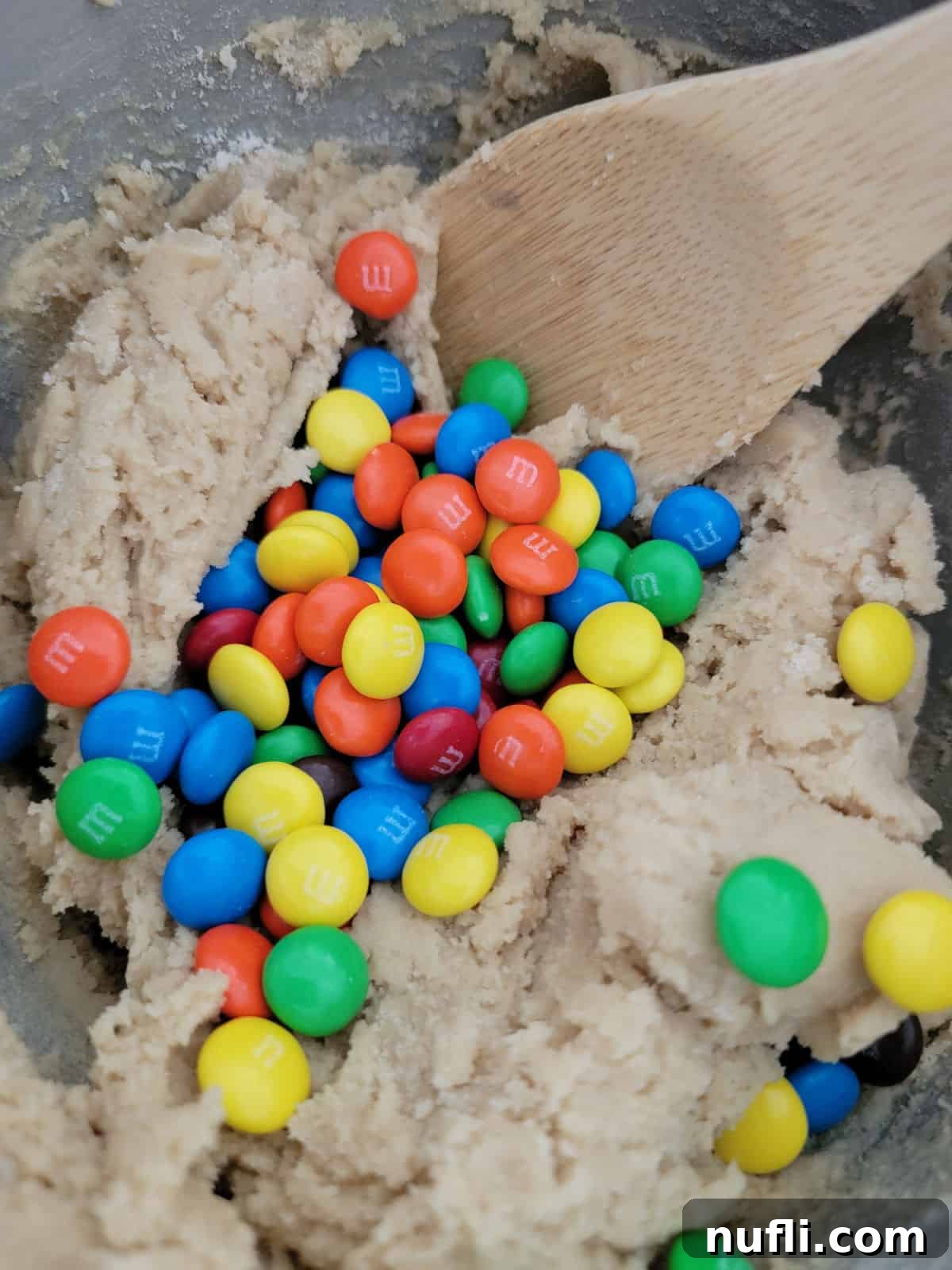 M&Ms folded into cookie dough with a wooden spoon in a mixing bowl.