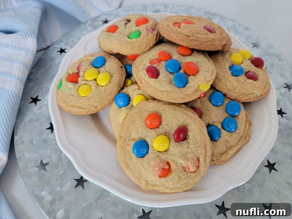 Colorful M&M Cookies arranged on a white plate on a silver platter, beside a cloth napkin.