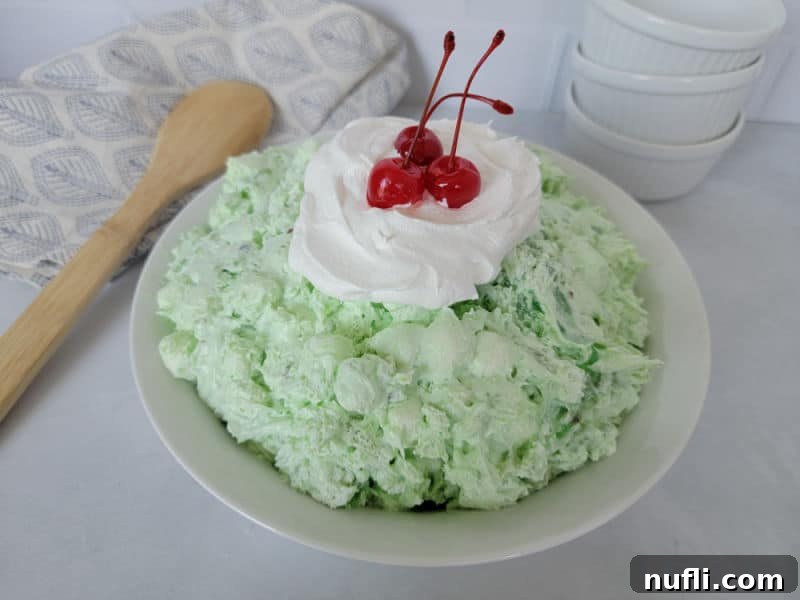 Classic Watergate Salad in a white bowl with a dollop of Cool Whip and Maraschino Cherries next to a wooden spoon and three white bowls