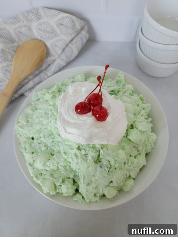Classic Watergate Salad in a white bowl with a dollop of Cool Whip and Maraschino Cherries next to a wooden spoon and three white bowls