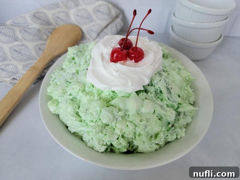 Easy Watergate Green Salad in a white bowl with a dollop of Cool Whip and Maraschino Cherries next to a wooden spoon and three white bowls