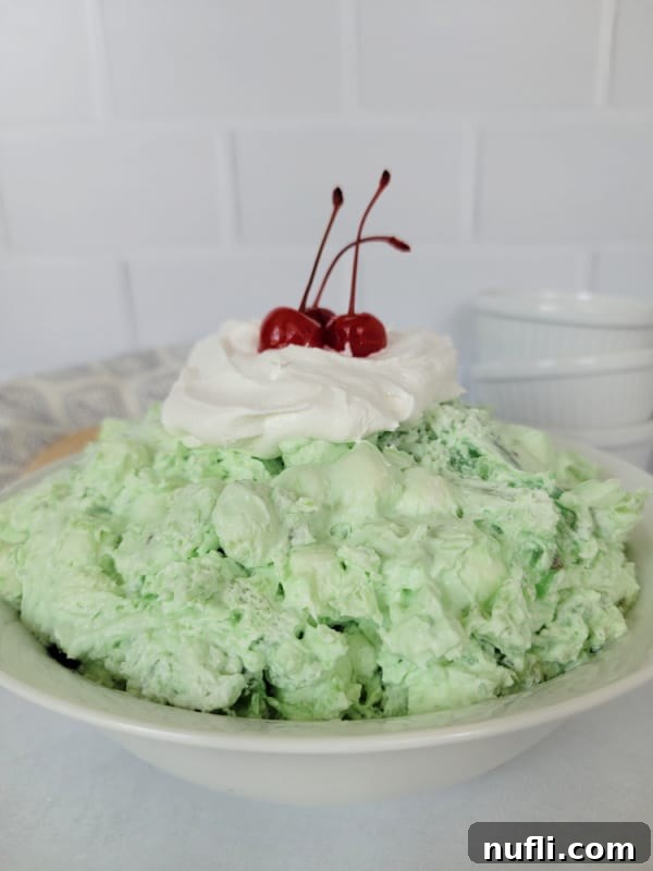 Classic Watergate Salad in a white bowl with a dollop of Cool Whip and Maraschino Cherries next to a wooden spoon and three white bowls