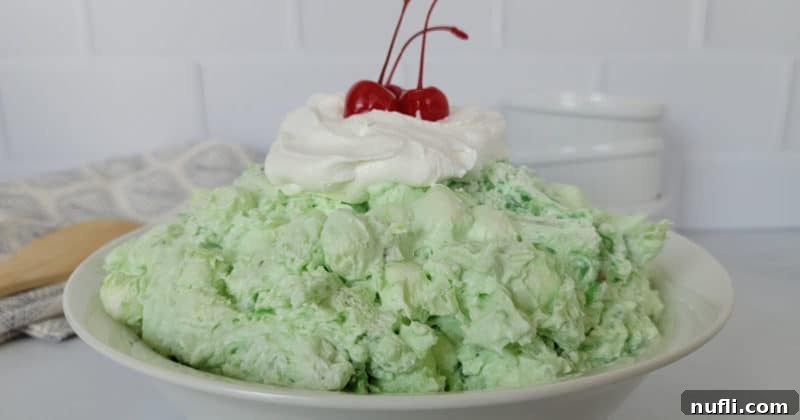 Classic Watergate Salad in a white bowl with a dollop of Cool Whip and Maraschino Cherries next to a wooden spoon and three white bowls