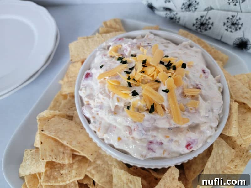 A beautifully garnished Fiesta dip in a white bowl, adorned with shredded cheese and surrounded by a generous scattering of tortilla chips, all presented on a elegant white platter next to a stack of plates.