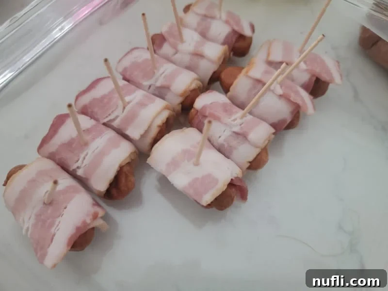 Unbaked bacon wrapped little smokies, each secured with a toothpick, arranged on a surface before coating.