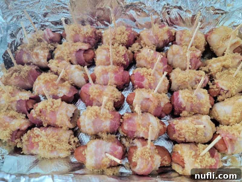 Neatly arranged rows of unbaked brown sugar bacon wrapped little smokies in an aluminum foil-lined baking dish, ready for the oven.