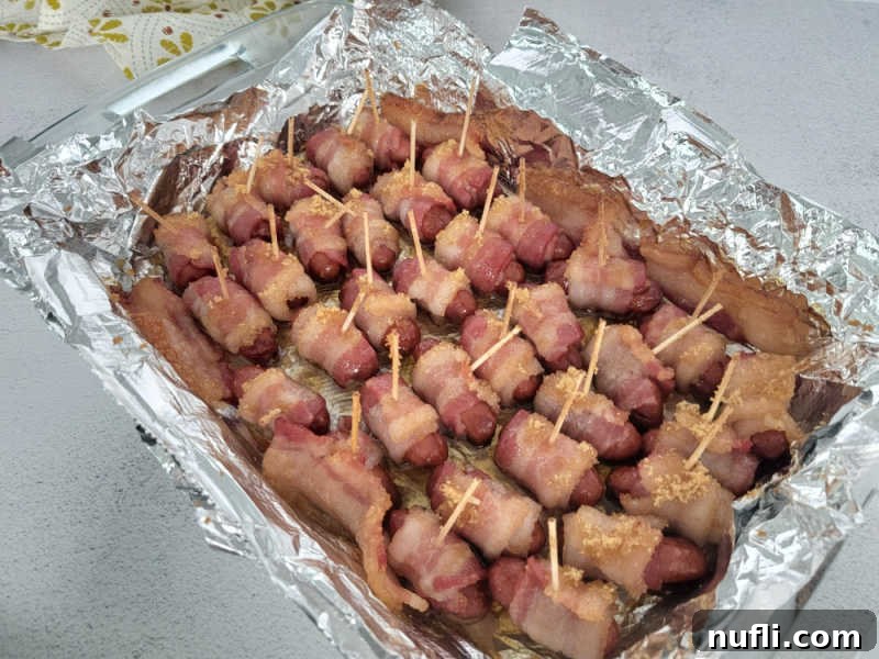 Freshly baked brown sugar bacon wrapped little smokies in an aluminum foil-lined baking dish, glistening with caramelized sugar.