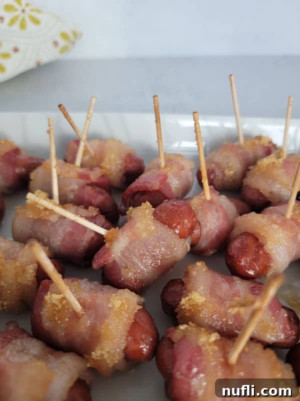 A single brown sugar bacon wrapped little smokie with a toothpick, glistening with glaze, resting on a white platter.
