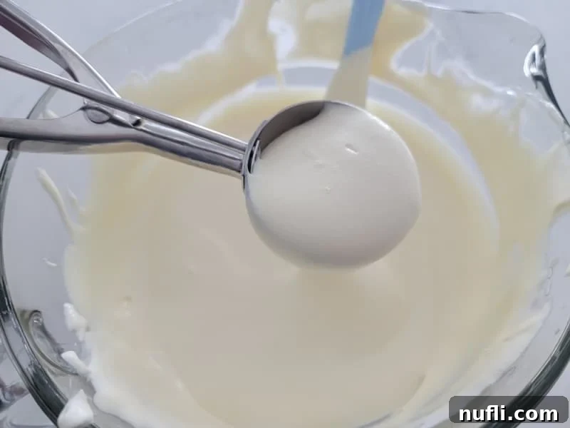 Scooping RumChata Pudding Shots mixture from a glass bowl into individual plastic cups using a cookie scoop