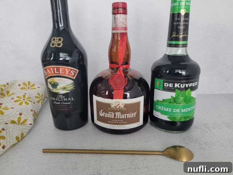 Bottles of Baileys Irish Cream, Grand Marnier, and Green Crème de Menthe, ready for mixing, next to a gold bar spoon and cloth napkin.