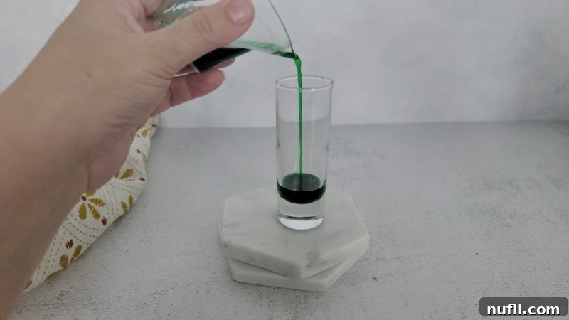 Green Crème de Menthe being poured into a shot glass on a white coaster, forming the first layer.