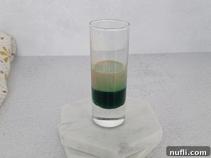 The second layer, Baileys Irish Cream, being carefully poured over a spoon onto the green layer in a shot glass.