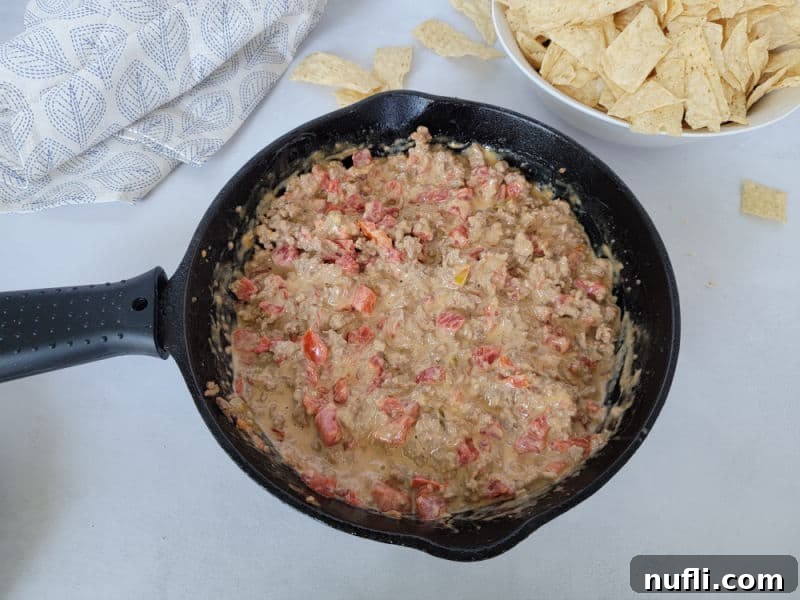The Easiest 3-Ingredient Velveeta Rotel Ground Beef Dip 9 A cast iron skillet filled with Velveeta Rotel Dip with ground beef, next to a white bowl overflowing with crunchy tortilla chips and a neatly folded cloth napkin.