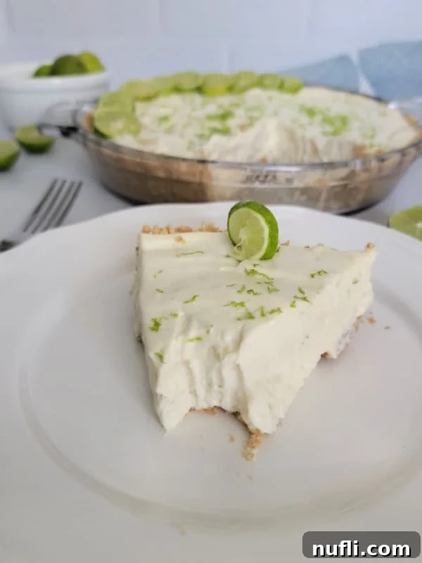 Slice of key lime pie with a bit taken of the front on a white plate next to a fork and the pie in the background. 